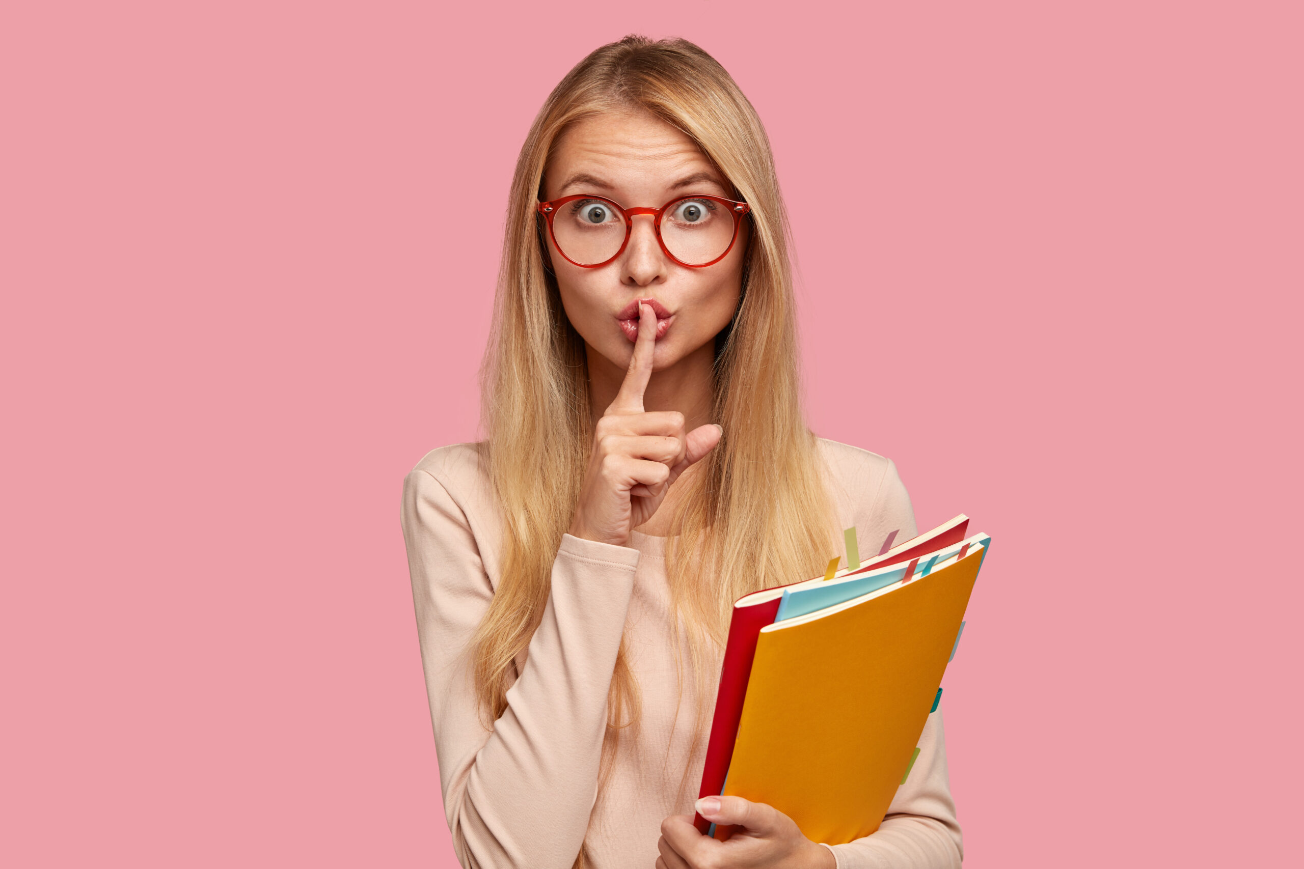 Omg, dont speak! Attractive Caucasian girl keeps index finger over lips, demonstrates silence gesture, asks not bother her, be quiet and speechless, wears spectacles with red rim isolated on pink wall